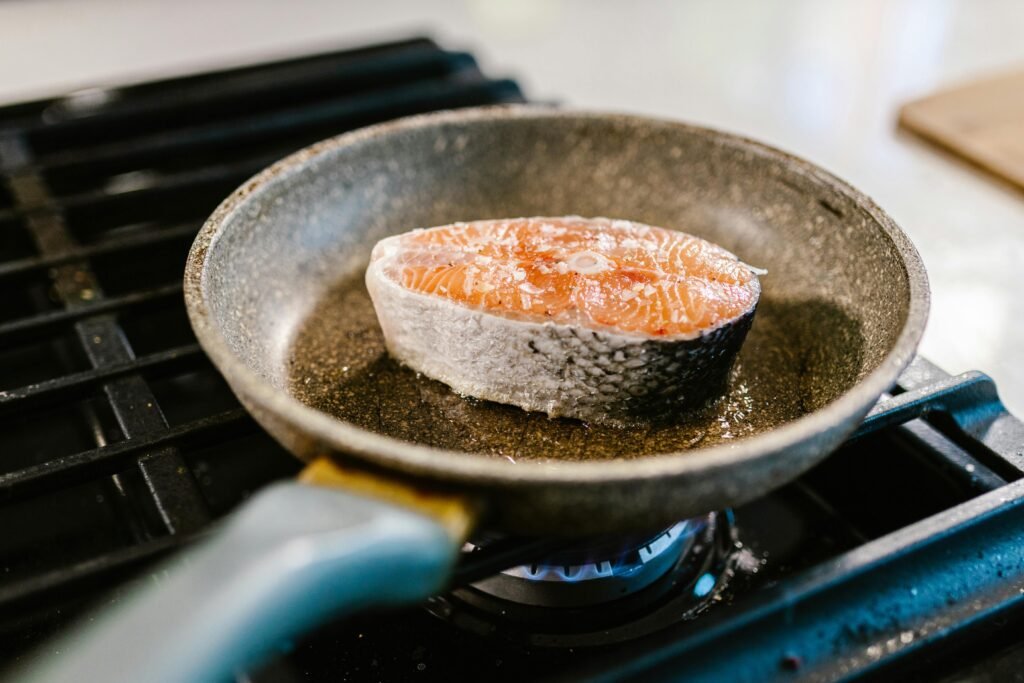 A salmon steak is searing in an iron frying pan on a stove, showcasing cooking detail.