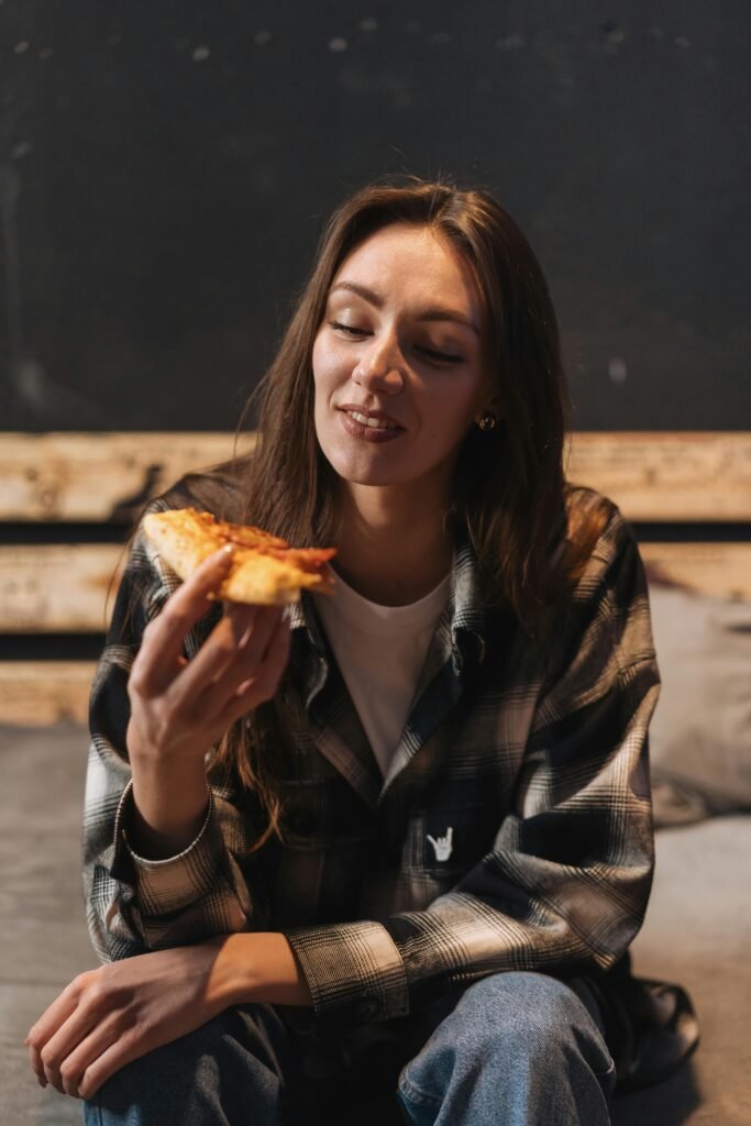 A woman sits on a couch indoors, holding and enjoying a slice of pizza with a rustic background.
