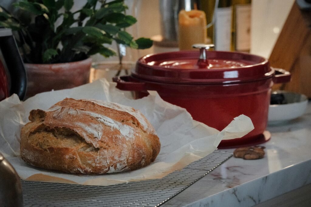 Close-up of a rustic loaf of bread on a kitchen counter with a red Dutch oven nearby.